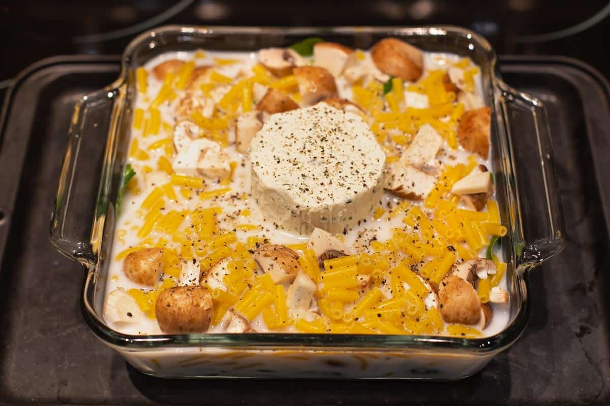 Uncooked pasta and mushrooms in a baking dish topped with a block of Boursin cheese.