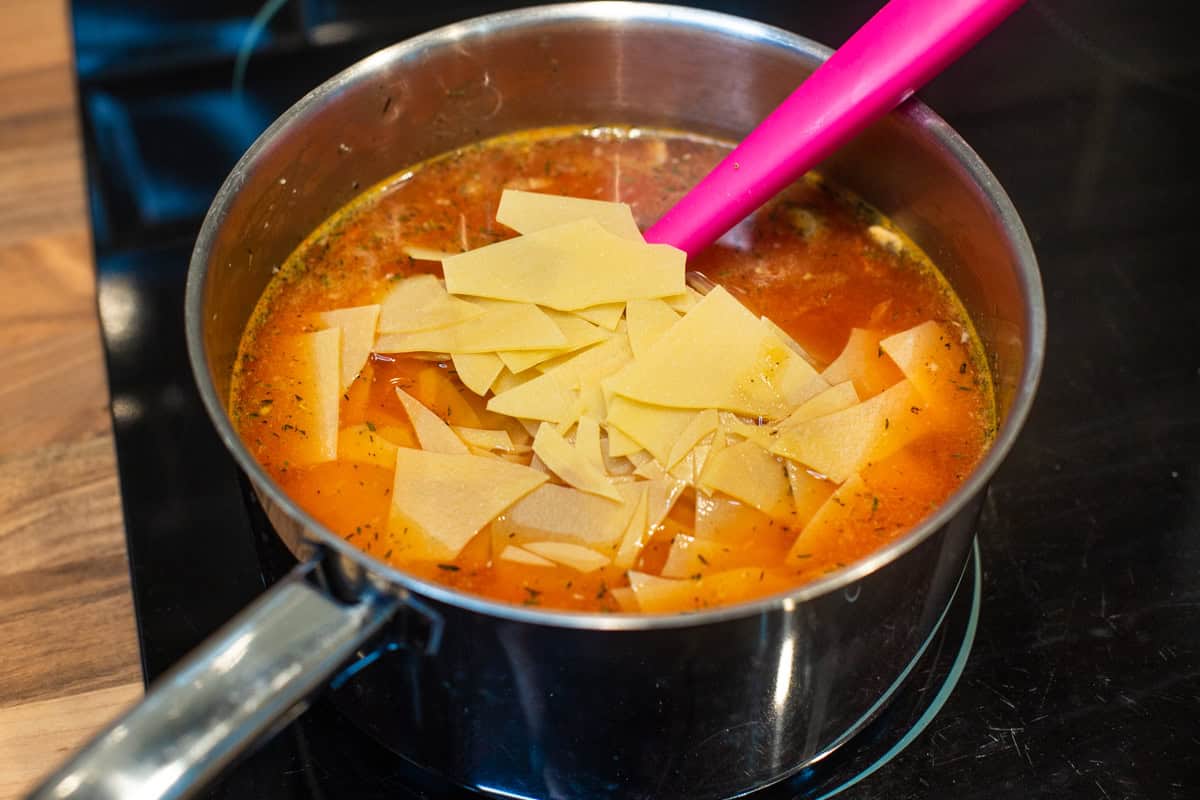 Broken pieces of uncooked lasagne on top of vegetable soup in a saucepan.
