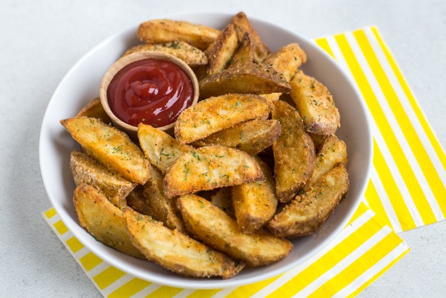 Crispy potato wedges in a bowl with a pot of ketchup and striped yellow napkins