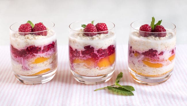 Three glasses of layered overnight oats with peaches and raspberry sauce, topped with fresh raspberries