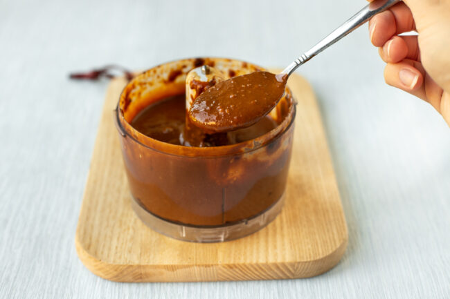 Peanut sauce in a food processor bowl being scooped with a spoon.