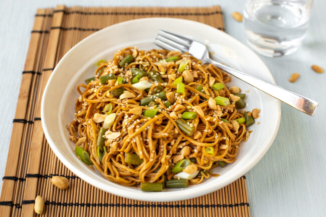 A bowlful of creamy peanut butter noodles topped with green veg and nuts.