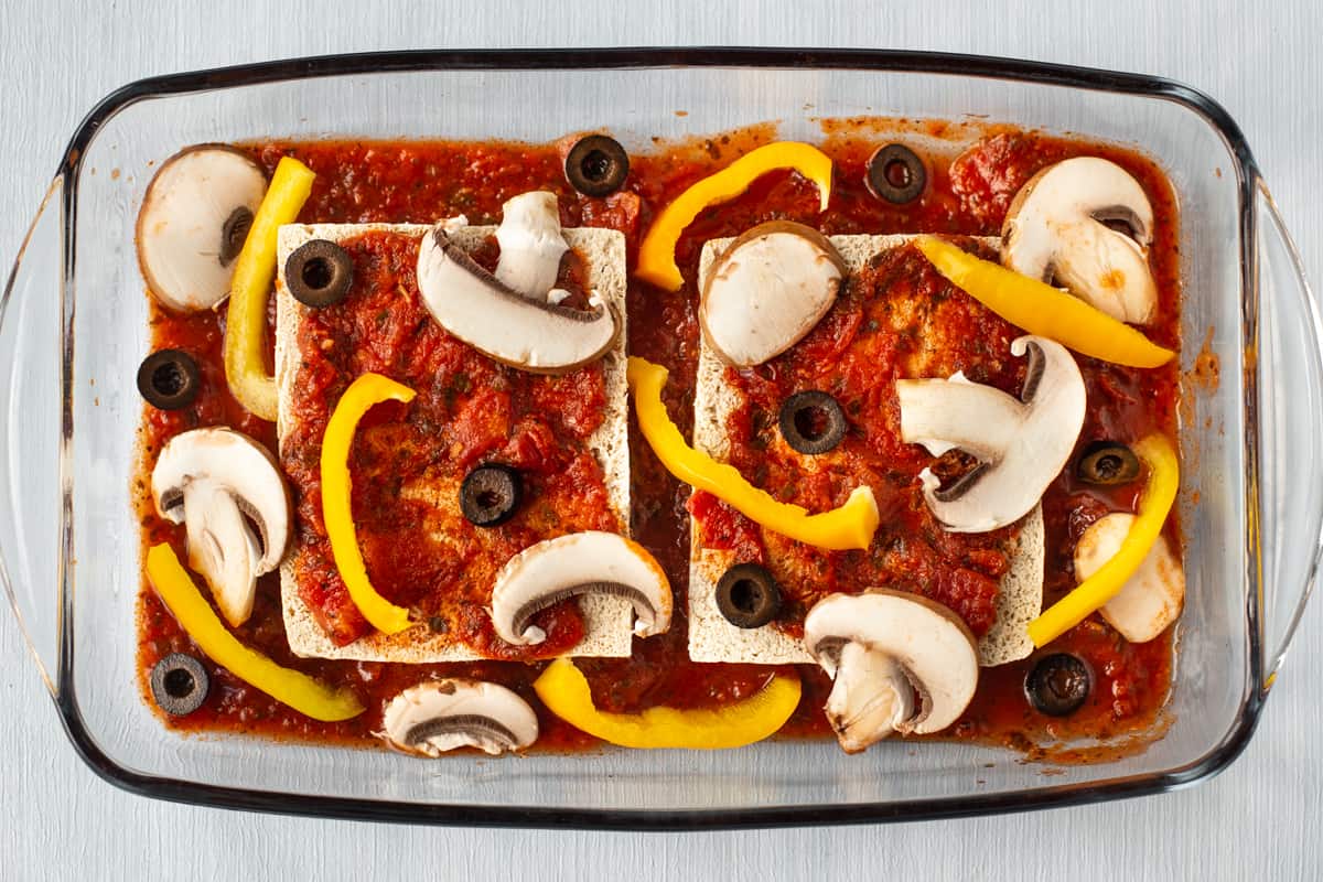 Tofu and tomato sauce in a baking dish, topped with raw vegetables.