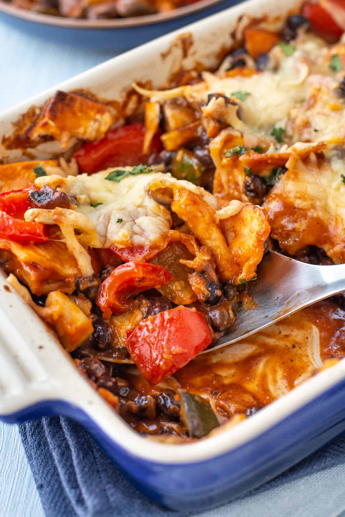 Close-up of a spoon scooping an enchilada casserole, with roasted vegetables and black beans.