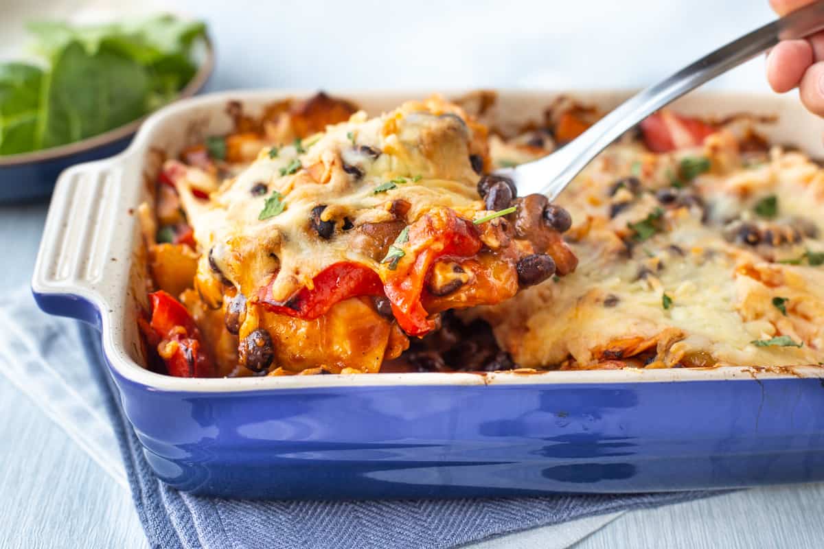 A large spoon taking a scoop of enchilada casserole from a blue baking dish.