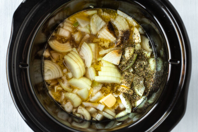 Raw onions and herbs in a slow cooker pot.