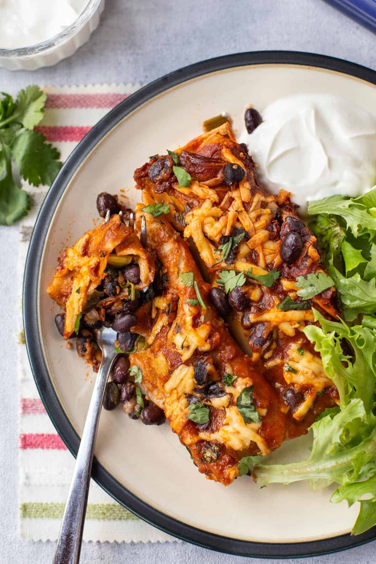 A fork cutting into black bean enchiladas with sour cream.