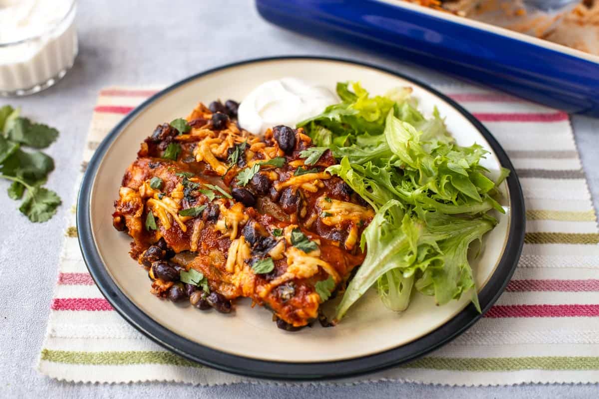 A portion of black bean enchiladas on a plate with salad and sour cream.