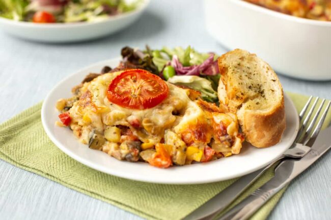 A portion of cheesy vegetarian lasagne on a plate with garlic bread and salad.