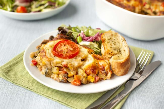 A portion of vegetable lasagne on a plate with garlic bread and salad.