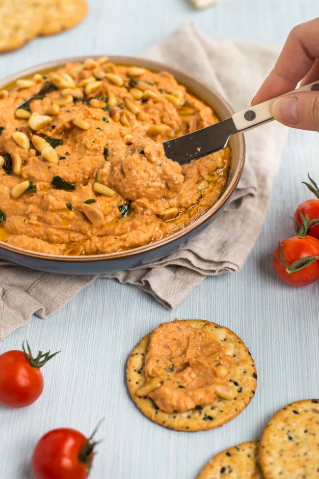 A knife taking a scoop of tomato hummus to spread on a cracker.