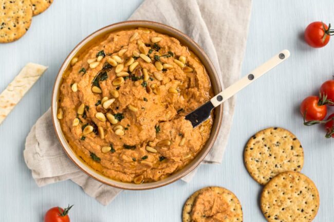 A knife taking a scoop of sun-dried tomato hummus topped with toasted pine nuts.