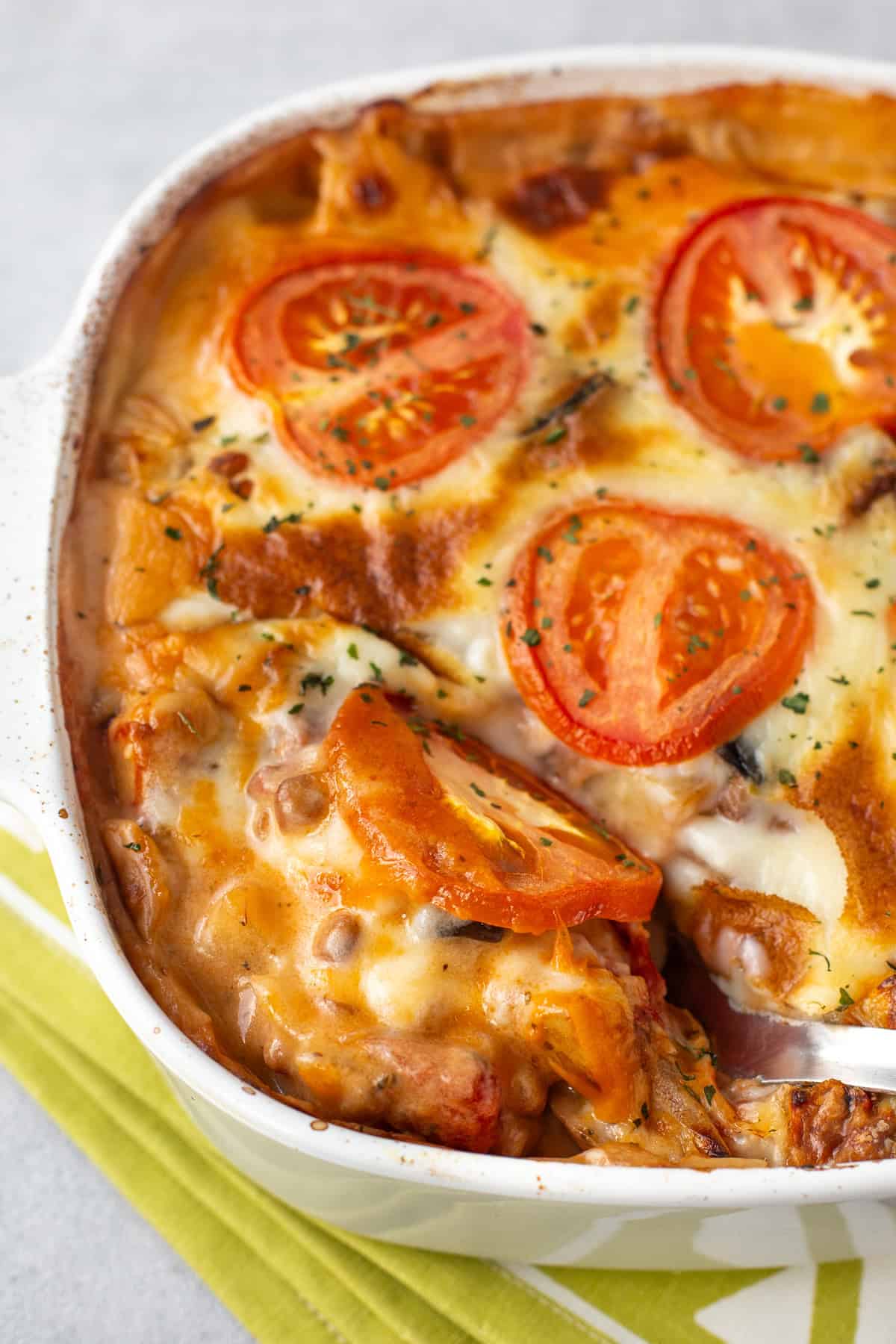 A spoon taking a scoop from a baking dish of cheesy moussaka casserole.