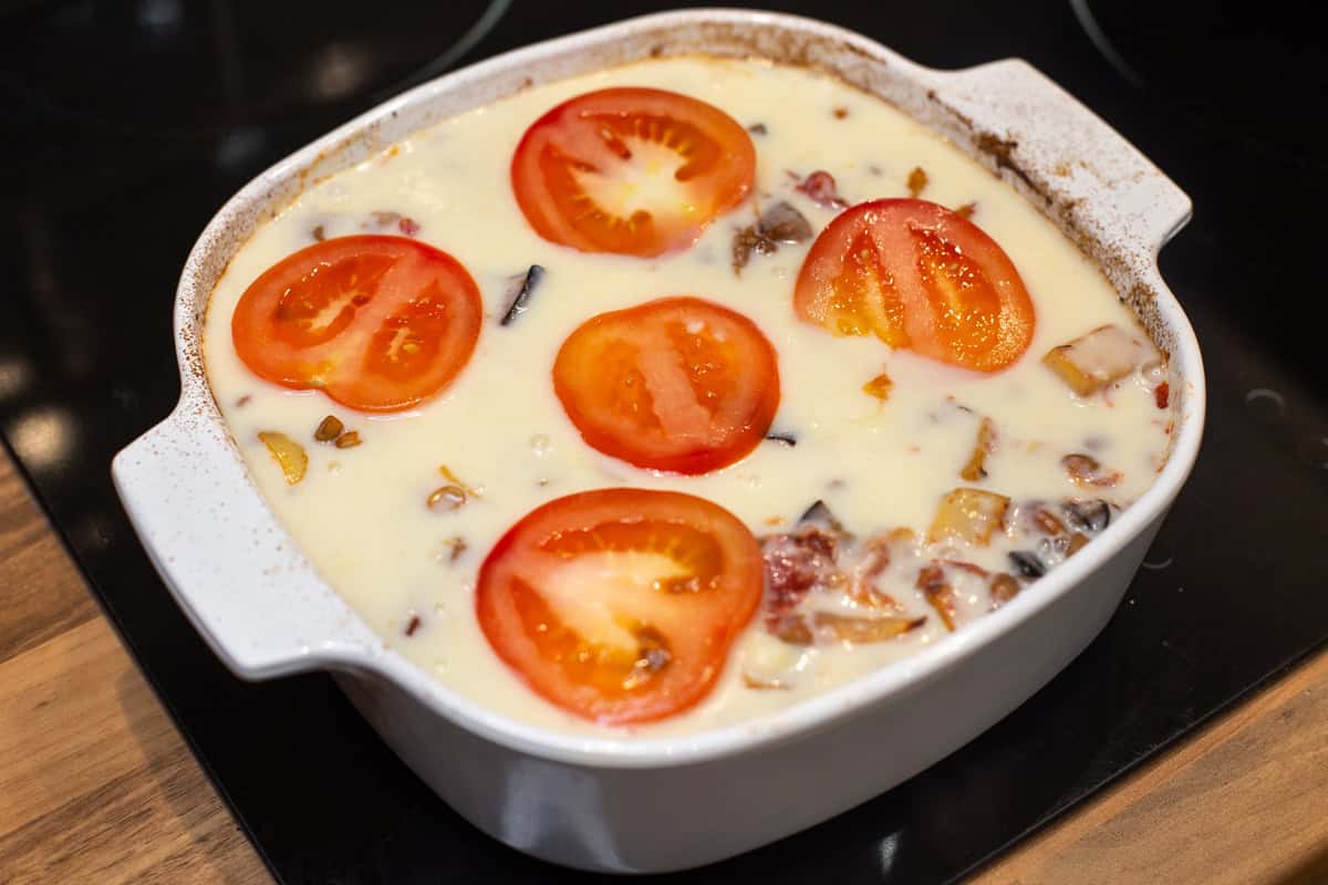 Uncooked moussaka in a baking dish topped with sliced tomatoes.
