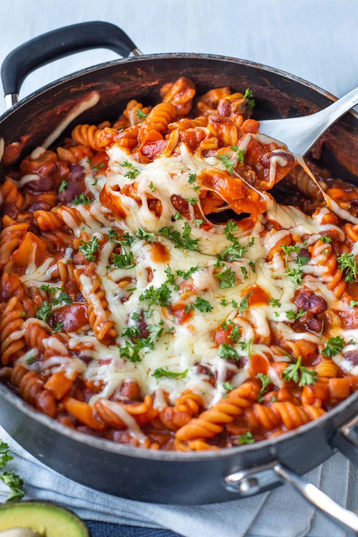 A spoon scooping one pot vegetarian chilli mac from a pan topped with gooey cheese.