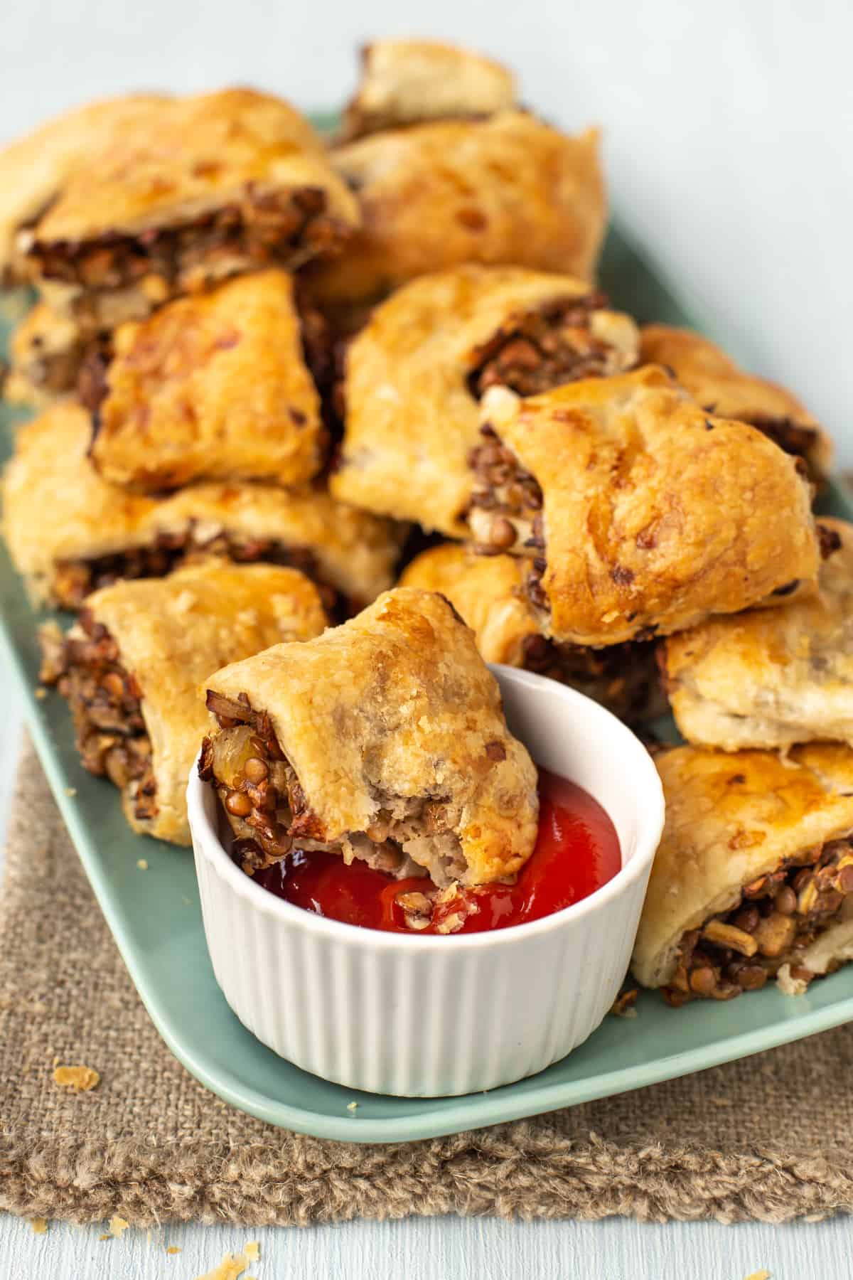 A lentil sausage roll resting on the edge of a pot of ketchup, with more vegetarian sausage rolls behind.