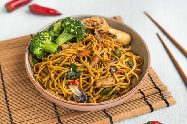 Veggie chow mein in a bowl with a spring roll and steamed broccoli.