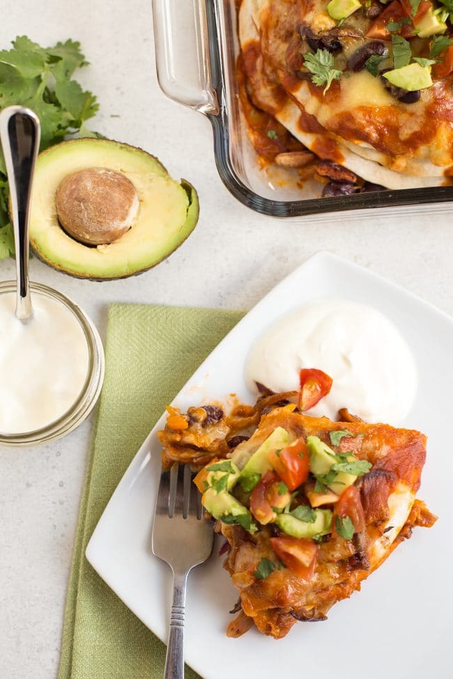 Portion of veggie enchilada stack on a plate with avocado and sour cream, shot from above