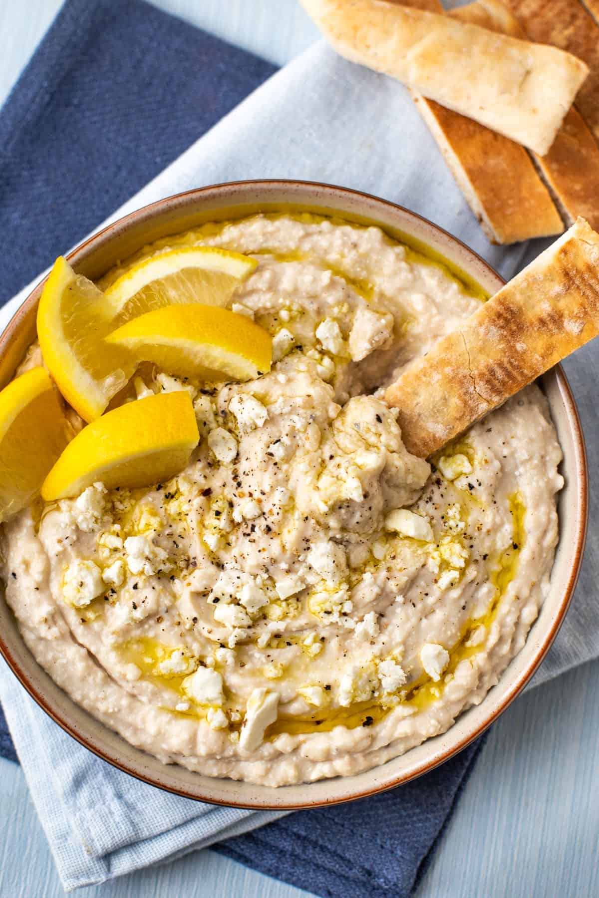 White bean hummus topped with lemon wedges, being scooped by a stick of pitta bread.